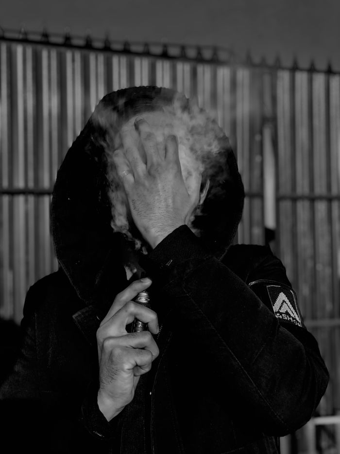 Black and white portrait of a hooded person vaping, obscured by smoke.